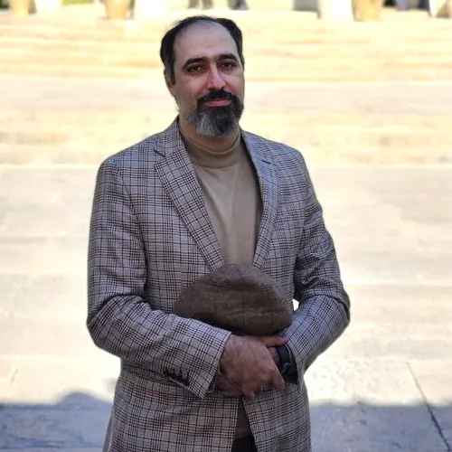 دکتر رضوی‌فرد ایستاده در آرامگاه حافظ شیراز، با لباس رسمی و پس‌زمینه گنبد و ستون‌های باغ حافظ.