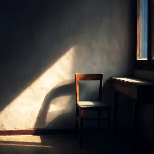 صندلی چوبی خالی کنار میز ساده، در فضایی کم‌نور با سایه‌های تیزِ نور پنجره روی دیوار.