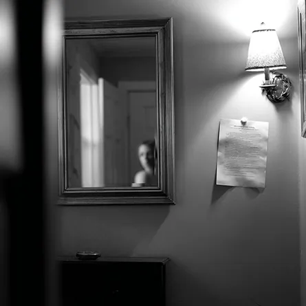 اتاقی کم‌نور با آینه‌ای که چهره فردی را در قاب بازتاب می‌دهد و برگه‌ای نصب‌شده بر دیوار؛ نمادی از تأمل، سکوت و بازخوانی درونی روایت انسان