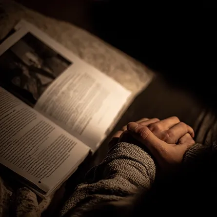 دست‌های گره‌خورده فردی در کنار کتابی باز در نور نرم شب؛ تصویری از خلوت اندیشه، تردید و جست‌وجوی معنا در لحظه‌ای انسانی
